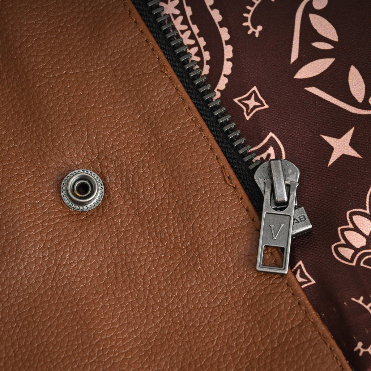 Close-up of the motorcycle club vest's metal zipper pull and front snap beside cognac cowhide leather and brown paisley lining.