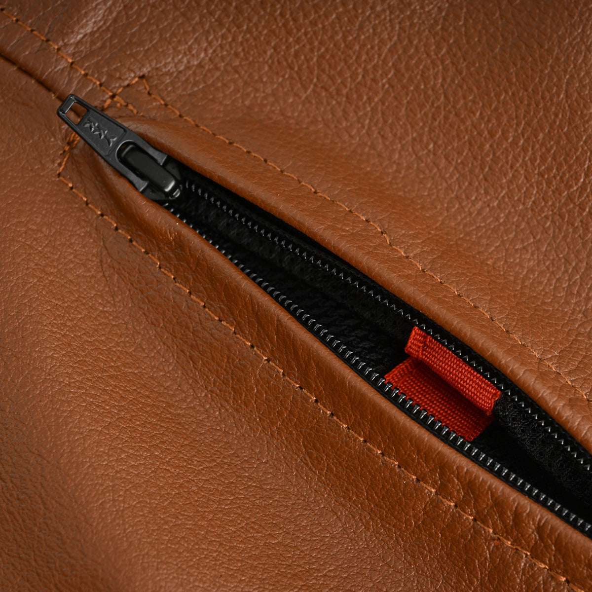 Open YKK zippered vent in cognac cowhide leather, showing black mesh and a red fabric tab inside the motorcycle jacket.