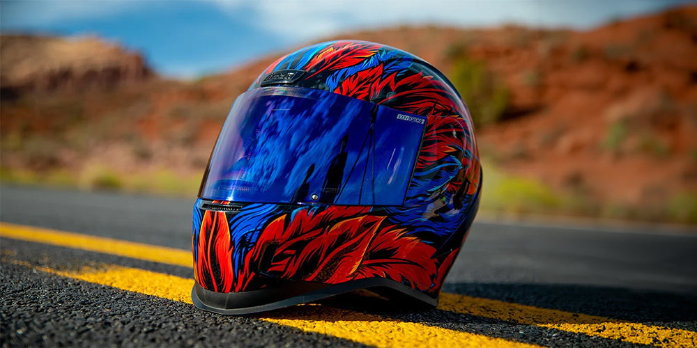 A full-face motorcycle helmet landing page image with a vibrant blue and red flame pattern resting on a paved road with a blurred desert landscape in the background. 