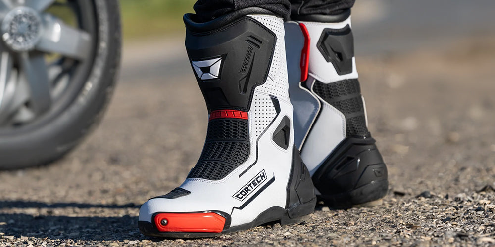 Motorcycle Boots Collection Landing Page Background Image, a close-up shot of a rider's feet wearing Cortech motorcycle boots on an asphalt surface, with a blurred motorcycle wheel in the background.
